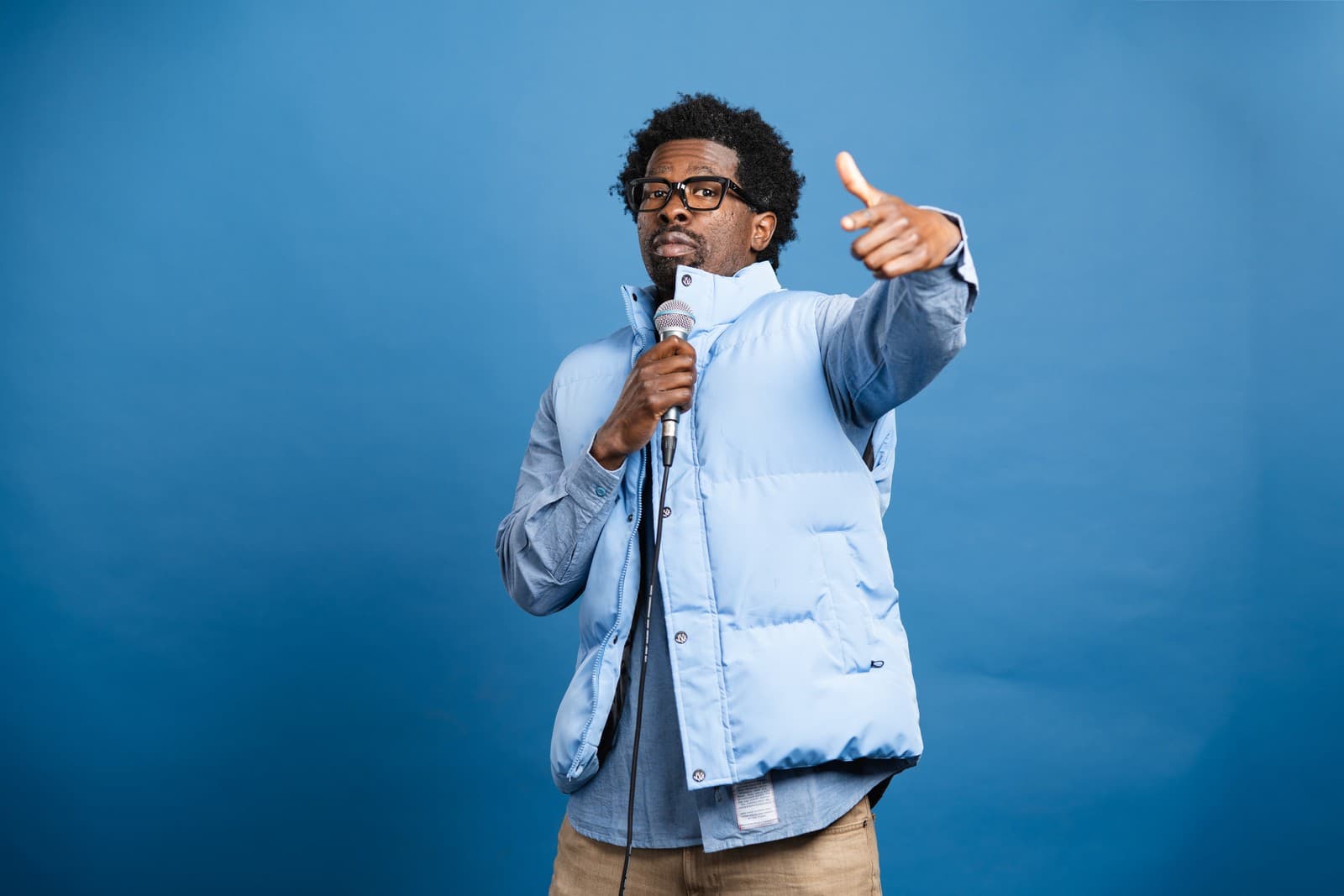 Stroy Moyd in a light blue vest, microphone in one hand, pointing toward the camera against a blue backdrop.
